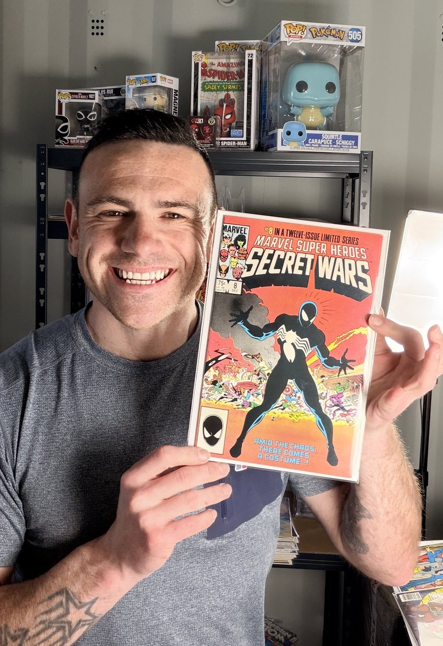 Lee from Retro Relics Comics holding a copy of Marvel Super Heroes Secret Wars #8, with his comic shelves and collection behind him